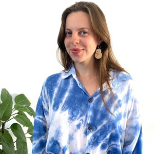 Puede incluir: Una mujer con el pelo largo y castaño lleva una camisa azul y blanca de tie-dye con cuello abotonado. Lleva unos pendientes grandes, redondos y de madera.
