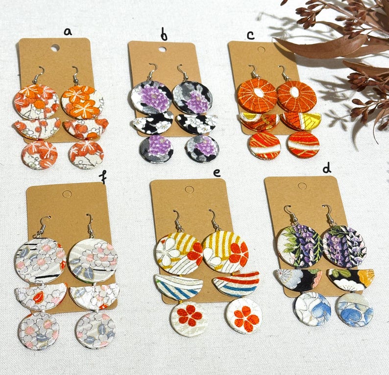 May include: Six pairs of earrings with three tiers of fabric circles. The fabric is patterned with flowers, geometric shapes, and abstract designs. The earrings are in various colors, including purple, orange, red, blue, and white.