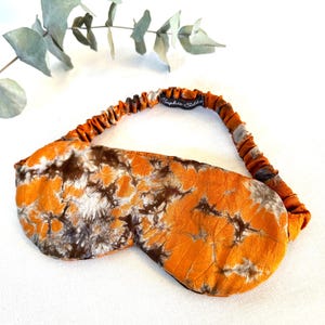 May include: A sleep mask featuring a tie-dye pattern in shades of orange, brown, and white. The mask has an elasticated strap with a black fabric label that reads Sophie Silks in white script text. The fabric has a soft, slightly wrinkled texture.