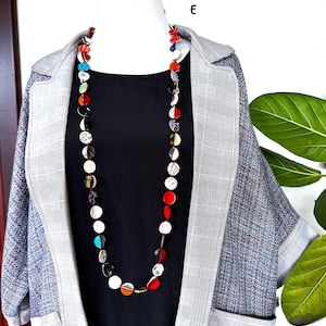 May include: A long necklace featuring a series of small, circular, multi-colored discs in shades of red, white, black, blue, and orange, strung together on a thin cord. The necklace is displayed on a mannequin wearing a black top and a grey plaid blazer.