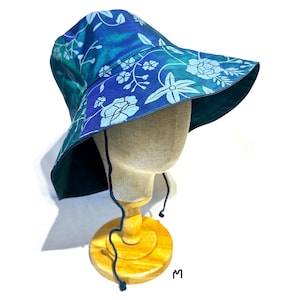May include: A wide-brimmed bucket hat featuring a vibrant iridescent blue and green fabric with a light blue floral pattern. The hat includes a dark blue chin strap with adjustable toggles, providing a secure fit for this colorful headwear.