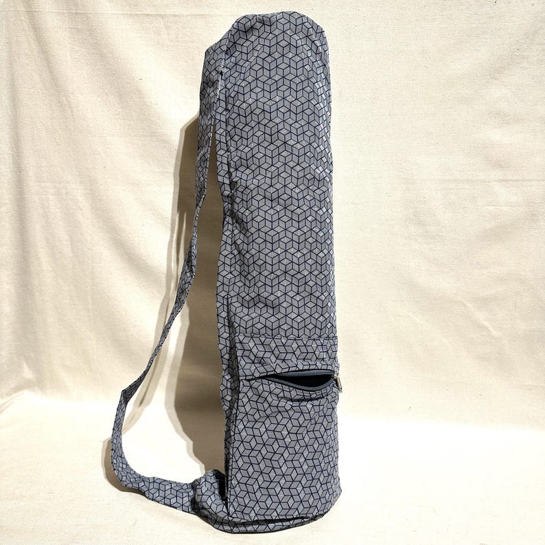 May include: A grey yoga mat bag featuring a geometric cube pattern. The bag has a zipped pocket and an adjustable shoulder strap. The bag is made of fabric and is designed to carry a yoga mat. The bag is approximately 76 cm long.