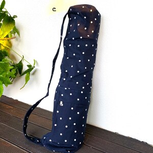May include: Navy blue yoga mat bag with white polka dots. The bag has a shoulder strap and a small pocket with a zipper. The bag is made of fabric and is designed to carry a yoga mat. The bag is leaning against a white wall.