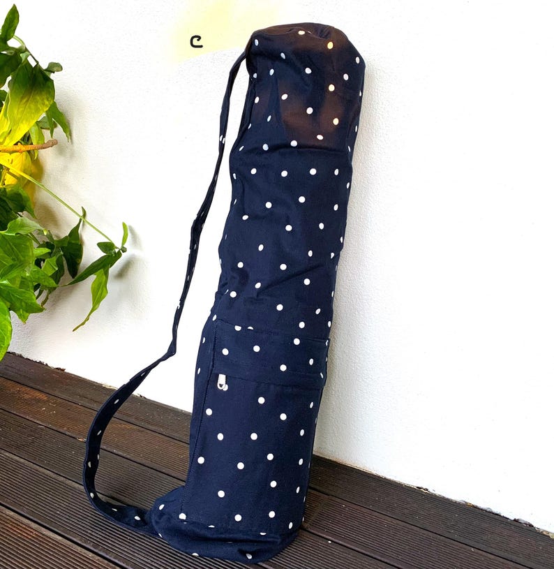 May include: Navy blue yoga mat bag with white polka dots. The bag has a shoulder strap and a small pocket with a zipper. The bag is made of fabric and is designed to carry a yoga mat. The bag is leaning against a white wall.