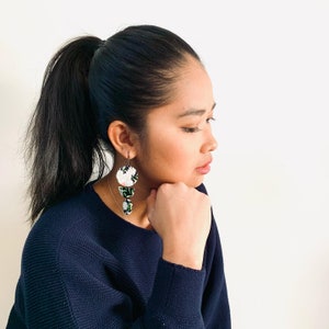 May include: A woman with long dark hair pulled back in a ponytail is wearing a dark blue sweater. She is wearing a pair of floral earrings with white and green accents.