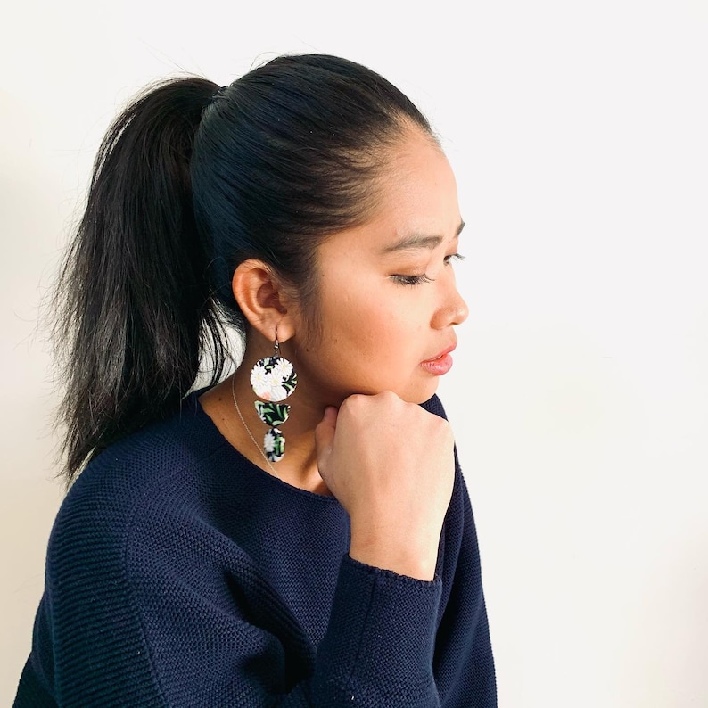 May include: A woman with long dark hair pulled back in a ponytail is wearing a dark blue sweater. She is wearing a pair of floral earrings with white and green accents.