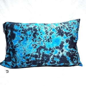 Könnte beinhalten: Ein rechteckiges Zierkissen mit einem lebendigen Batikmuster in den Farben leuchtendes Cyan, elektrisches Blau und tiefes Schwarz. Der Stoff zeigt ein komplexes, organisches, netzartiges Design auf der gesamten Oberfläche. Der Buchstabe D ist schwarz aufgedruckt.
