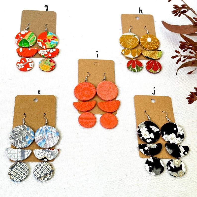 May include: Assortment of handmade earrings featuring colorful patterned fabric circles and half-circles. The earrings are displayed on brown card stock with letters 'g', 'h', 'i', 'j', and 'k' printed on them.