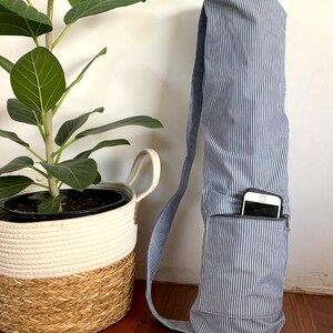 May include: A blue and white striped yoga mat bag with a shoulder strap and a zipped pocket. A smartphone is visible in the pocket. A potted plant and a woven basket are in the background.