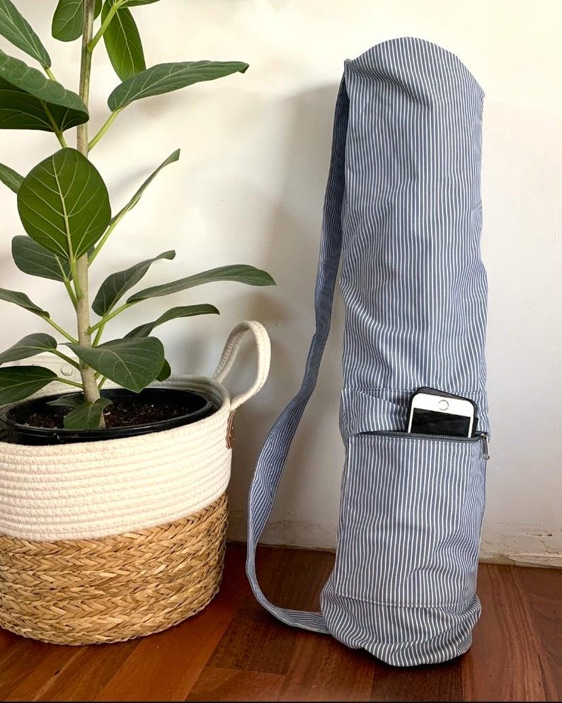 May include: A blue and white striped yoga mat bag with a shoulder strap and a zipped pocket. A smartphone is visible in the pocket. A potted plant and a woven basket are in the background.