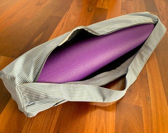 Puede incluir: Una esterilla de yoga morada es parcialmente visible dentro de una bolsa de yoga a rayas azules y blancas. La bolsa tiene cierre de cremallera y una correa para el hombro. La bolsa y la esterilla están sobre una superficie de madera.