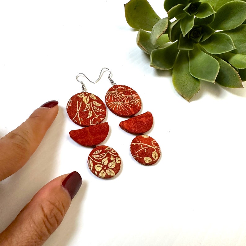 May include: A pair of red and gold earrings with a geometric design. The earrings are made of fabric with a floral pattern and are suspended from silver hooks.