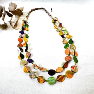 May include: A double-strand necklace featuring numerous small, circular, flat discs in a variety of colors and patterns, including orange, green, yellow, purple, and striped designs, strung on a thin green cord with a braided fabric neck section.