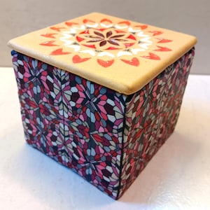 May include: A small square box with a lid. The box is decorated with a geometric pattern of pink, red, and black. The lid is a light brown color and has a white and red floral design.