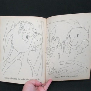 May include: A black and white illustration of a rabbit painting a round, smiling character. The text "There. Now, just a minute." is below the illustration. The illustration is from a children's book.