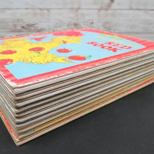 May include: A stack of vintage children's books with a red, white, and blue cover. The cover features a yellow chick with red apples. The title of the book is "The Red Book".