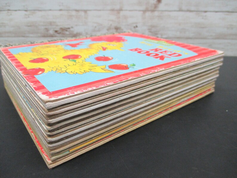 May include: A stack of vintage children's books with a red, white, and blue cover. The cover features a yellow chick with red apples. The title of the book is "The Red Book".