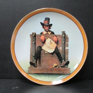 May include: Decorative plate featuring a historical scene. The image depicts a man in a wooden stockade holding a sign that reads "For Gluttony." The plate has a gold rim with a decorative border.
