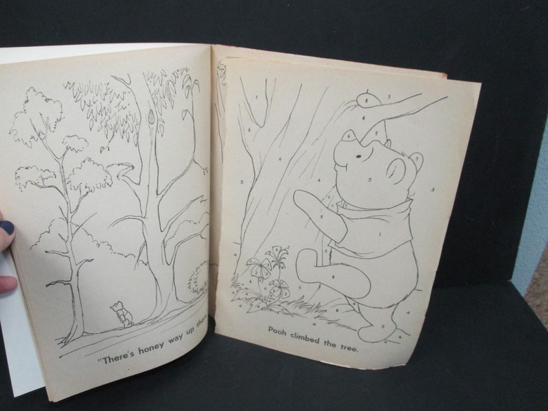 May include: A black and white colouring book page featuring Winnie the Pooh climbing a tree. The page has numbers on it for a connect-the-dots activity. The text on the page reads "Pooh climbed the tree."