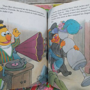 May include: Bert, a yellow Sesame Street character, plays a record on a vintage record player with a large, purple horn. Ernie, another Sesame Street character, dances with a snowman wearing a blue hat and a black jacket.