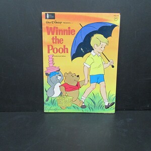 May include: A vintage Winnie the Pooh book cover featuring Winnie the Pooh, Piglet, and Christopher Robin. The cover is yellow with a blue umbrella and green grass. The title "Winnie the Pooh" is in red and black lettering.
