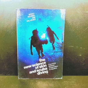May include: A book cover with a blue background featuring two scuba divers in silhouette. The title reads "The New Science of Skin and Scuba Diving." Text at the top says "Newly Revised and Enlarged."