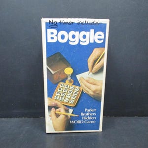 May include: A white box with the word "Boggle" in large blue letters. The box shows a person writing on a piece of paper and a game board with letter tiles. The text on the box says "Parker Brothers Hidden Word Game".