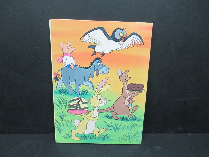 May include: A colourful illustration featuring Winnie the Pooh characters: Piglet riding on Eeyore, Rabbit carrying a stack of sandwiches, Kanga holding a cake, and Owl flying overhead. The background is a sunset orange with green grass.