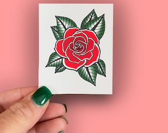 May include: A red rose with green leaves, drawn in a traditional tattoo style. The rose is the main focus of the image, with the leaves surrounding it.