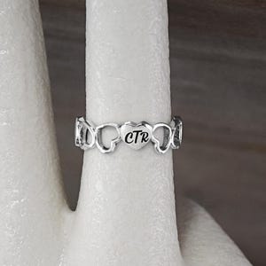 May include: A silver ring with a heart-shaped design. The heart has the initials "CTR" engraved inside.