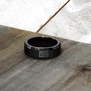 May include: A black ring with a geometric design and the letters "CTR" engraved on the side. The ring is set on a wooden surface, showcasing its dark color and modern style. The ring's design is angular and bold.