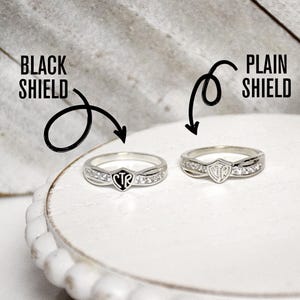 May include: Two silver rings with a shield design. The shield on the left ring is black with the letters CTR inside. The shield on the right ring is plain silver with the letters CTR inside. Both rings have small clear stones around the shield.