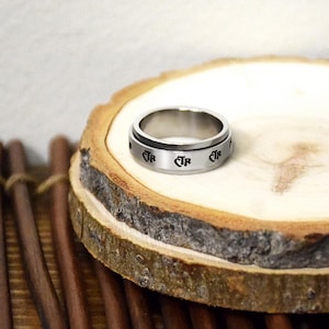 May include: A silver-colored spinner ring with the letters "JR" engraved around the band. The ring is displayed on a wooden slice, showcasing its metallic sheen and design.