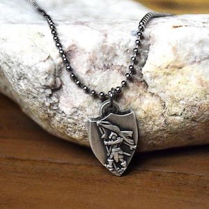 May include: A silver-toned pendant necklace featuring a shield-shaped charm. The charm depicts a detailed engraving of a figure holding a flag. The necklace chain is made of small, round beads. The pendant is displayed against a textured, light-colored stone.