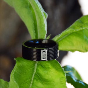 May include: A black ring with a grooved design and the letters "CTR" in a white rectangle. The ring is set against a backdrop of green leaves, creating a contrast of colors and textures. The ring appears to be made of metal.