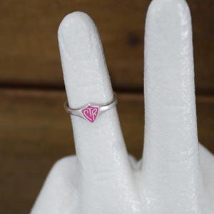 May include: A silver ring with a pink shield-shaped emblem. The emblem features the letters "CTR" in a stylized font. The ring is displayed on a white surface against a blurred brown background.