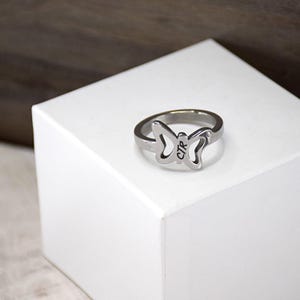 May include: A silver butterfly ring with the inscription "CTR" on the back. The ring is displayed on a white box.