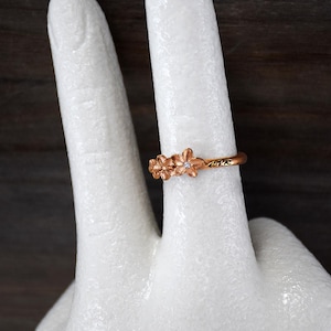May include: A rose gold ring featuring a floral design with three flowers. One flower has a small, clear gemstone in the center. The ring is on a white surface. The word "evr" is engraved on the band.