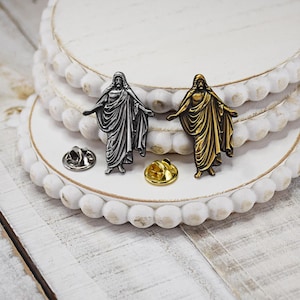 May include: Two lapel pins featuring a depiction of Jesus Christ. One pin is silver and the other is gold. Both pins are on a white background.