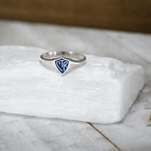 May include: A silver ring with a blue shield-shaped emblem. The emblem features the letters "STR" in a stylized font. The ring is set against a white, textured surface.