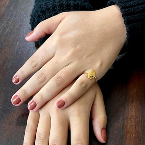 May include: A gold ring with a round, flat top featuring the engraved letters "CTR". The ring is worn on a finger with brown nail polish.