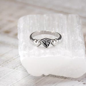 May include: A silver ring with a shield-shaped center featuring the letters "CTR" in black. Two heart-shaped accents flank the shield. The ring is displayed on a white, textured surface.