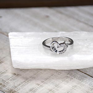 May include: A silver ring with a heart-shaped design and small clear stones. The word "CTR" is engraved on the ring.