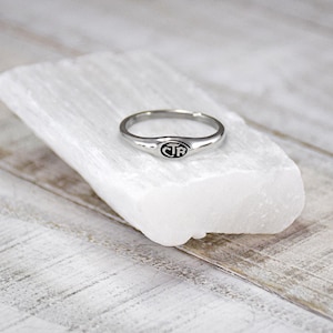 May include: A silver signet ring with a black oval-shaped face featuring the letters "CTR". The ring is displayed on a white, rectangular stone against a light wood-grain background. The ring is a simple band with a slightly raised face.