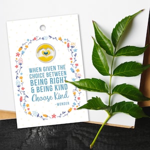 May include: A pin with a yellow and white heart design, set against a white card with the text "When given the choice between being right & being kind, Choose Kind." The card is decorated with a floral wreath. A green leafy branch is in the background.