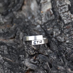 May include: A silver ring with the letters "CTR" engraved on the front. The ring is set against a dark, textured background, possibly wood or stone. The ring's design is simple and modern.
