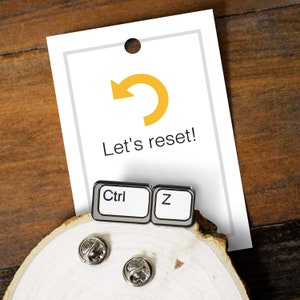 May include: A white and silver Ctrl Z pin, with the text "Ctrl" and "Z" on the keys, is displayed on a wooden surface. A white card with the text "Let's reset!" and a yellow curved arrow is behind the pin.