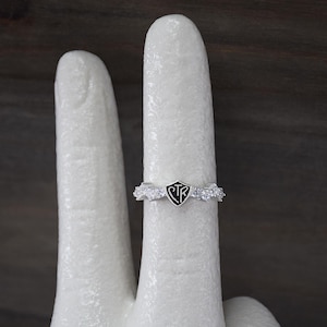 May include: A silver ring with a black shield-shaped emblem featuring the letters "CTR" in white. The ring is adorned with small, clear gemstones. The ring is displayed on a white surface.