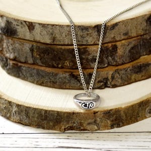 May include: Silver-toned necklace featuring a ring pendant with the letters "CTR" in a black oval. The chain is a delicate curb style. The pendant hangs against a backdrop of layered, textured wood slices.