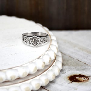 May include: Silver ring with a shield design in the center, featuring the letters "CTR". The ring has a black border and decorative patterns on the sides. The ring is displayed on a white, beaded surface.